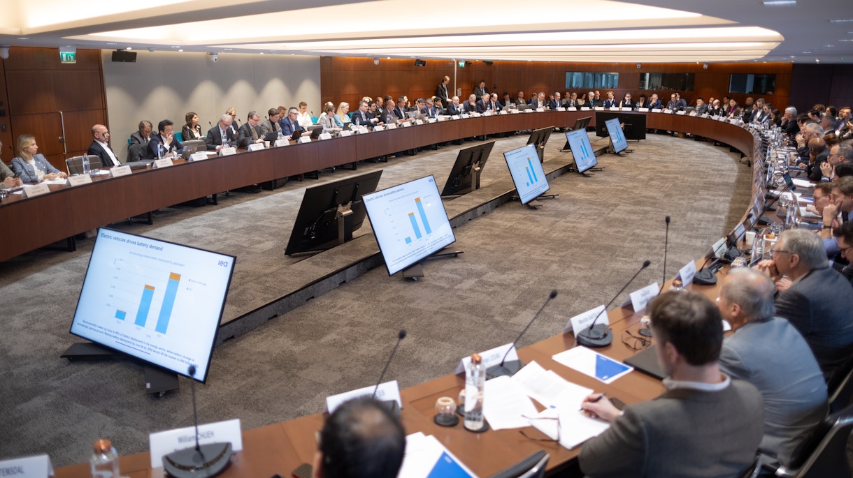 Image shows a large conference room with rounded tables across the edges with people sitting in the chairs